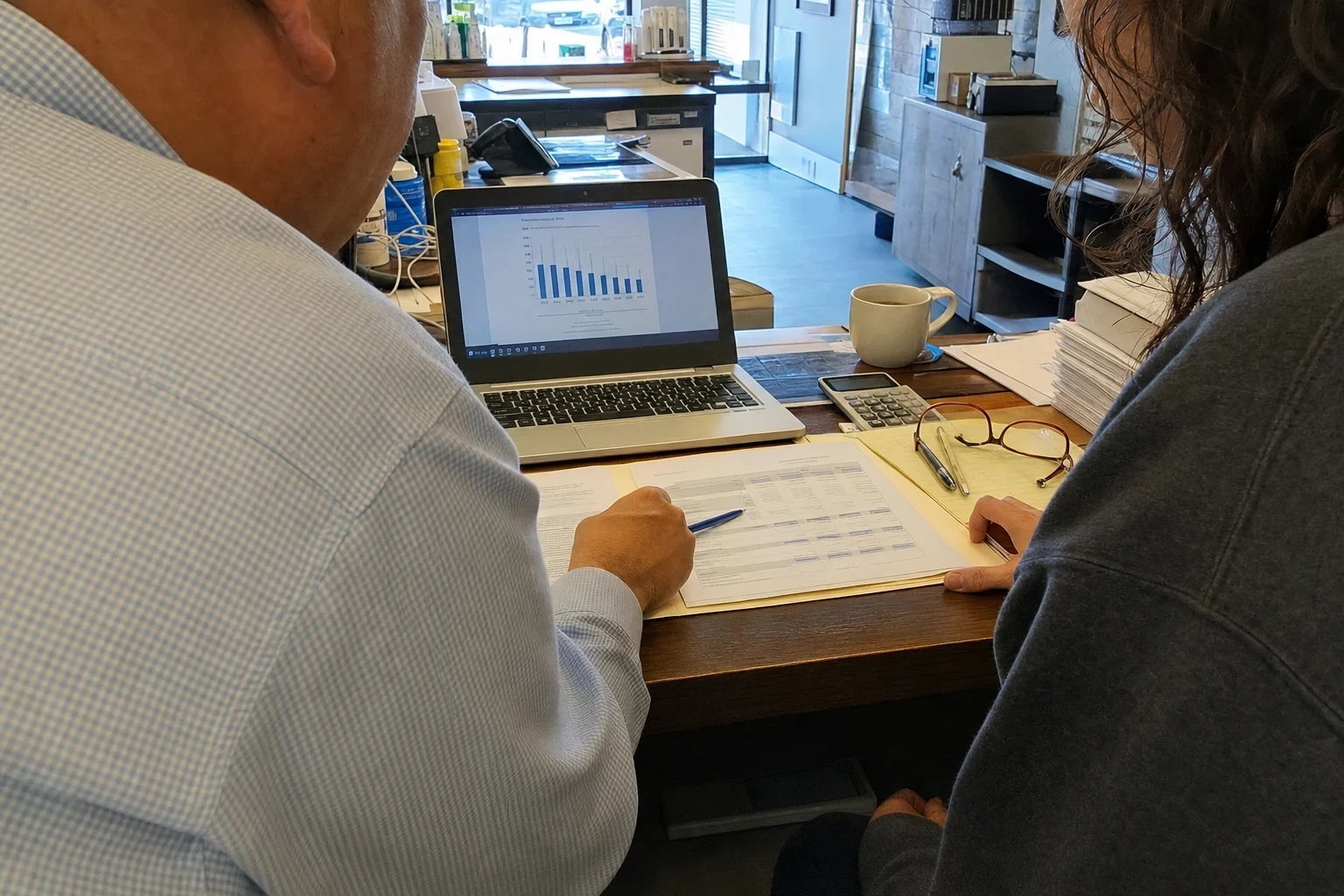 Over-the-shoulder view of two people at a cluttered shop desk reviewing a printed form, with a laptop showing a bar chart, a calculator, reading glasses and a coffee mug